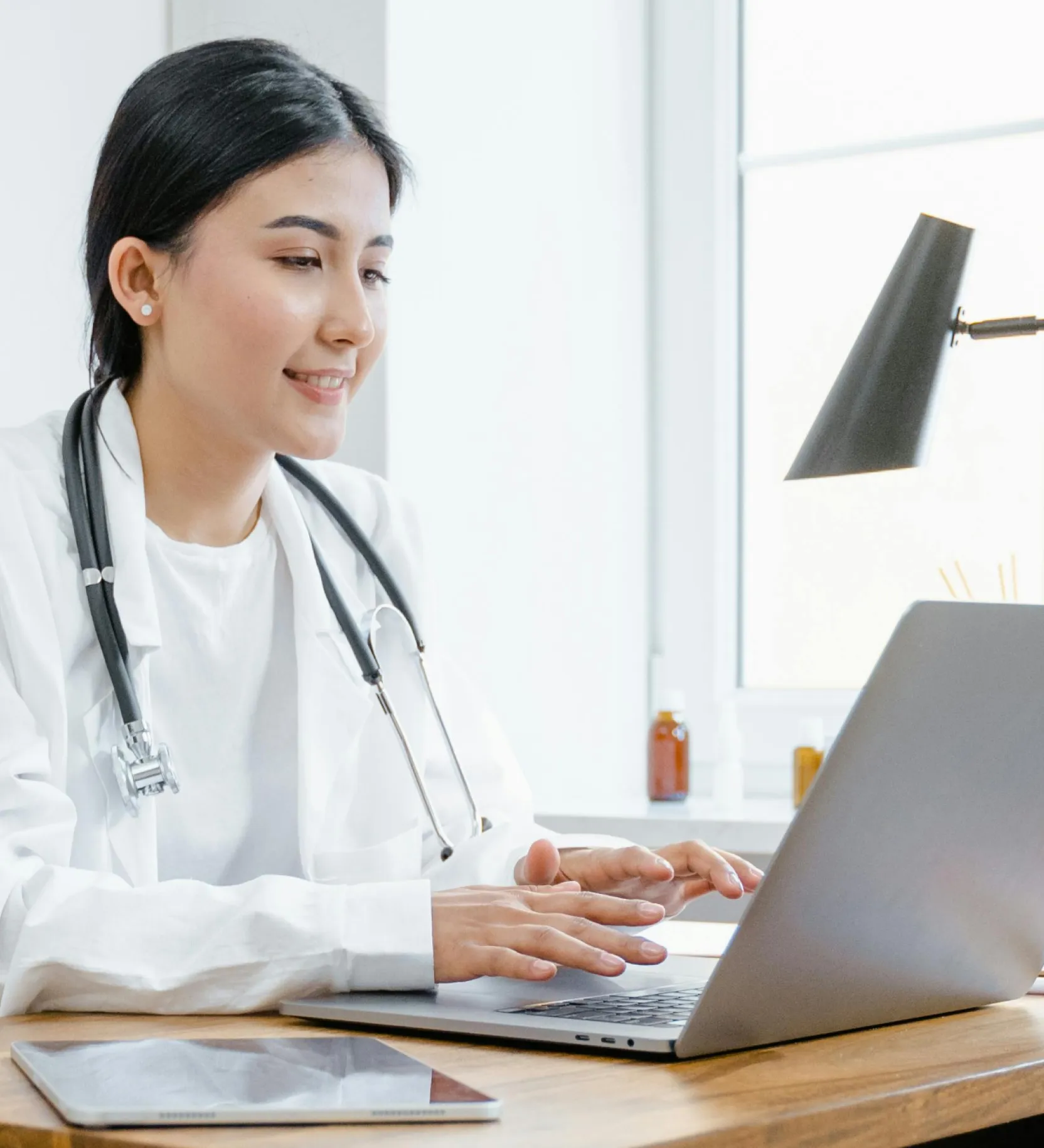 A woman in a white coat sits at a desk, focused on her laptop, surrounded by a professional workspace.