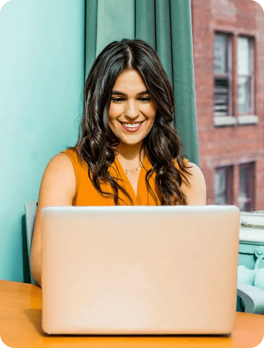 A woman smiles while working on a laptop, showcasing a positive and engaged expression.